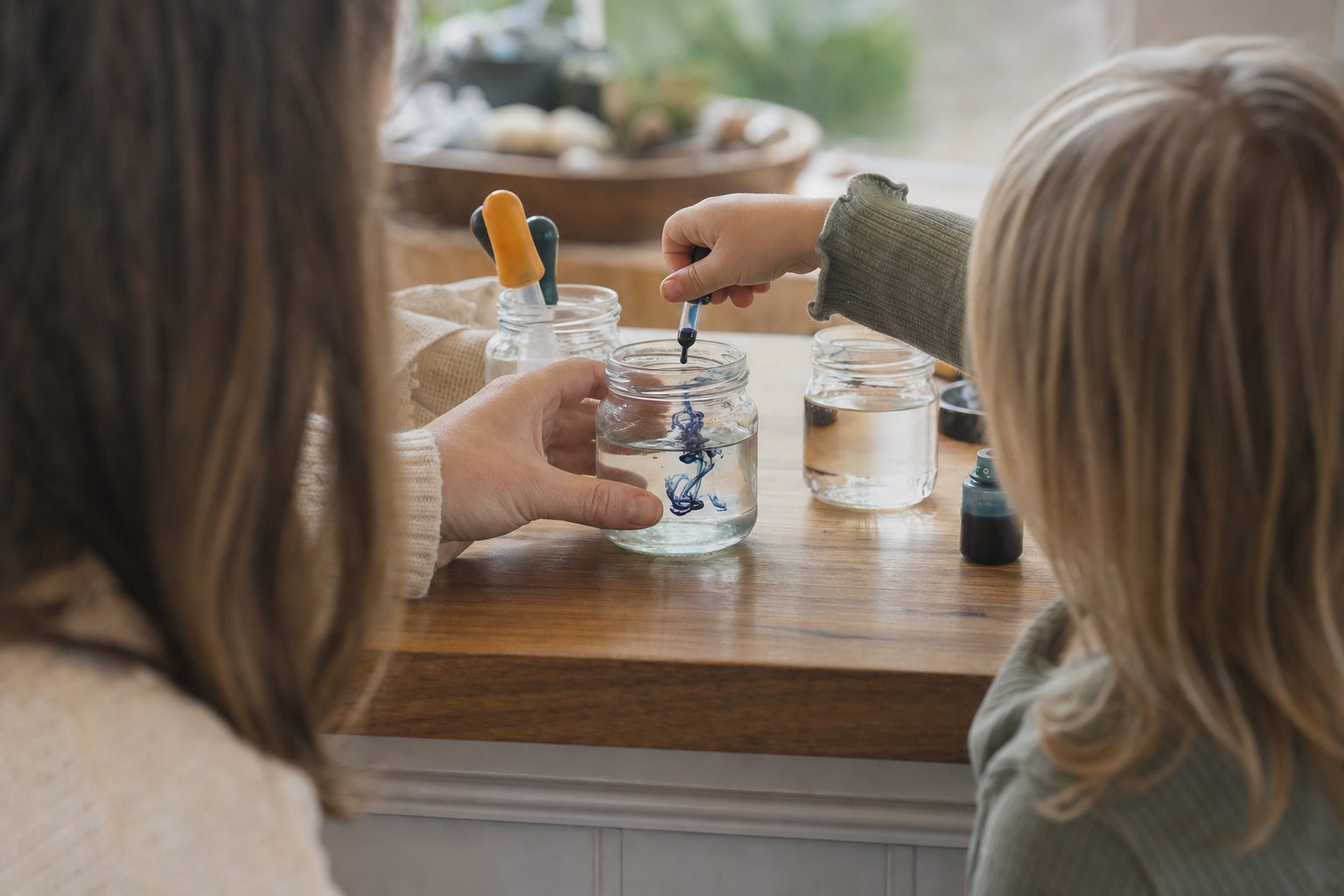 A parent and young child leaning in together over jars of coloured water at a wooden bench