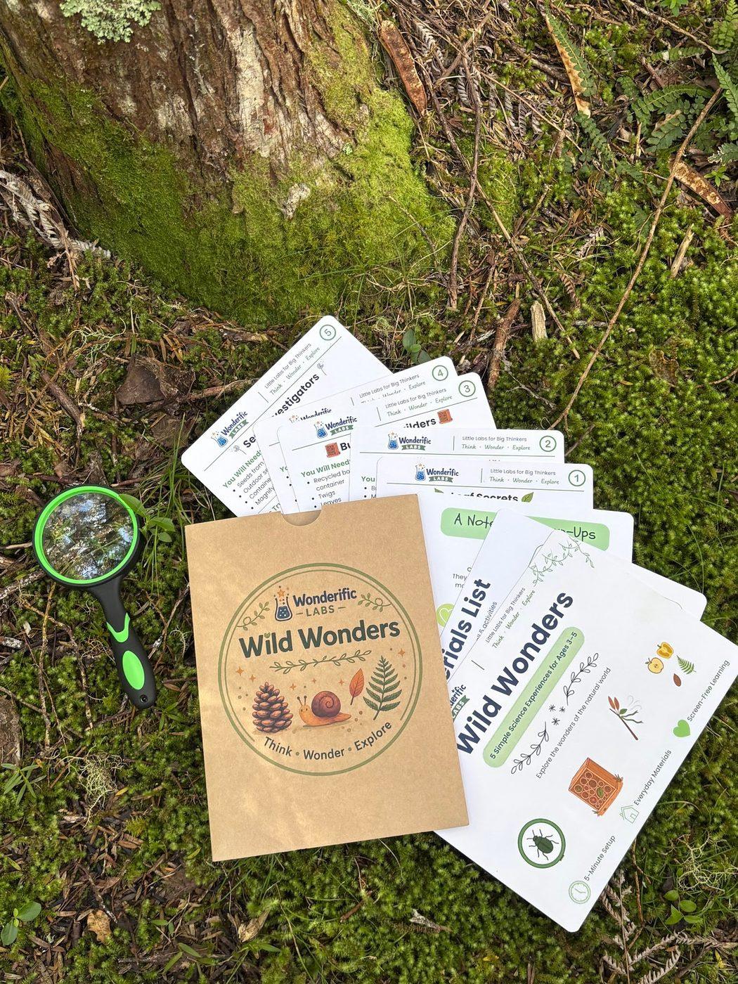 The Wild Wonders pack, activity cards and a magnifying glass laid out on moss at the base of a tree
