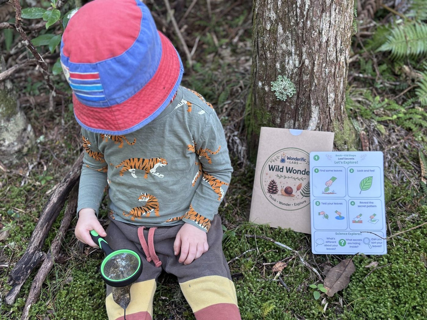 A child in a bucket hat crouching beside the Wild Wonders pack, holding a magnifying glass over a find