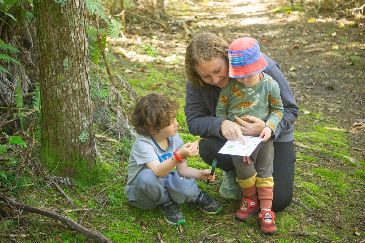 Dani sitting on the forest floor with two children, looking at an activity card together